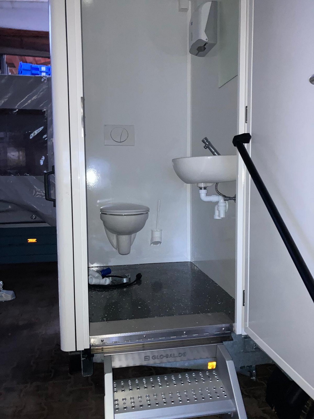 Das Bild zeigt den Innenraum einer mobilen Toilette. Links befindet sich eine Toilette, rechts ein kleines Waschbecken mit Wasserhahn. Der Boden ist fliesenartig, und eine Treppe führt zum Eingang. Die Wände sind einfach gehalten und in neutralen Farben.