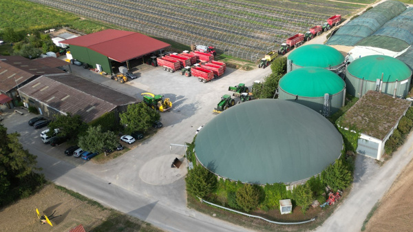 Luftaufnahme eines landwirtschaftlichen Betriebs. Im Vordergrund sind mehrere große, grüne Biogasanlagen und Lagerhallen sichtbar. Umgeben von Feldern und einem Weinbaugebiet. Straßen und Fahrzeuge sind ebenfalls zu sehen. Die Landschaft ist geprägt von landwirtschaftlicher Nutzung.