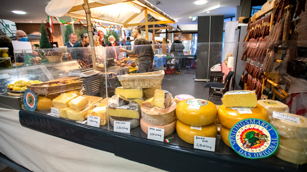 Ein Marktstand mit einer Auswahl an Käse. Verschiedene Käsesorten, die in Kästchen und Laiben präsentiert werden, sind gut sichtbar. Preise und Bezeichnungen sind an den Käsepackungen angebracht. Im Hintergrund sind Menschen zu sehen, die den Markt besuchen.