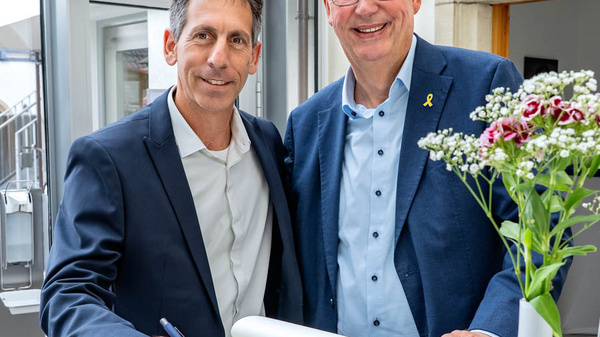 Zwei Männer stehen lächelnd zusammen an einem Tisch mit einem offenen Buch. Einer der Männer trägt einen Anzug, der andere ein Hemd und eine Jacke. Auf dem Tisch befindet sich ein Blumenstrauß. Im Hintergrund ist eine moderne Umgebung zu sehen.