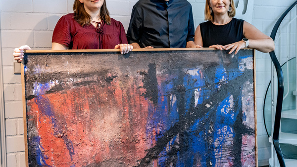 Drei Personen stehen nebeneinander und präsentieren ein großes, abstraktes Kunstwerk. Die Frau links trägt ein rotes Oberteil, der Mann in der Mitte ein schwarzes Hemd, und die Frau rechts hat ein weißes Oberteil an. Im Hintergrund ist eine geschwungene Treppe sichtbar.