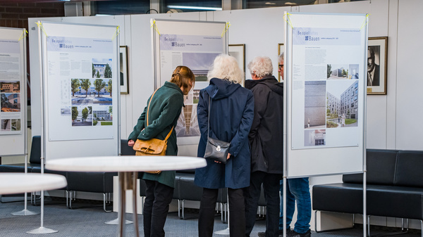 Vier Personen stehen umwis eine Ausstellungswand, die Informationen und Bilder präsentiert. Sie betrachten aufmerksam die Tafeln, die Gebäude und Landschaften zeigen. Im Hintergrund sind weitere Wandtafeln sichtbar, sowie Sitzgelegenheiten und Tische im Raum.