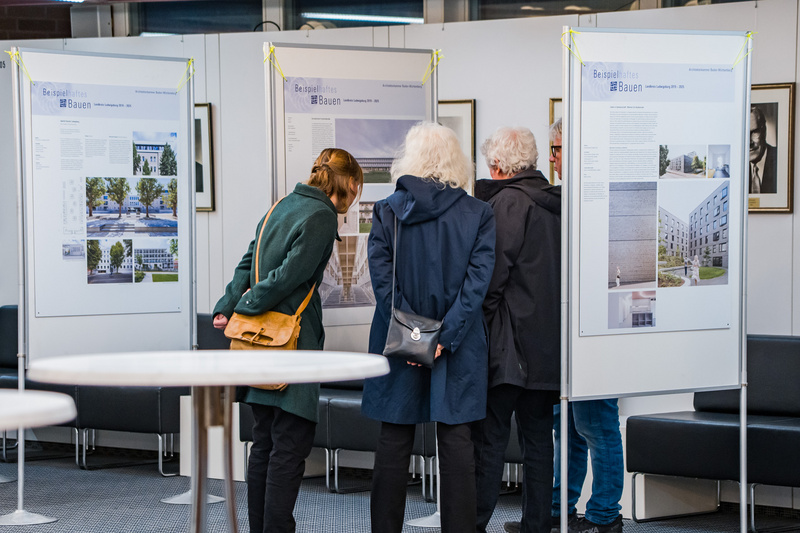 Vier Personen stehen umwis eine Ausstellungswand, die Informationen und Bilder präsentiert. Sie betrachten aufmerksam die Tafeln, die Gebäude und Landschaften zeigen. Im Hintergrund sind weitere Wandtafeln sichtbar, sowie Sitzgelegenheiten und Tische im Raum.