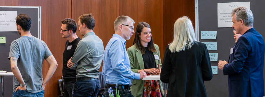 Header Bild: Eine Gruppe von sieben Personen steht in einem Konferenzraum und unterhält sich. Einige Notizen und Plakate sind im Hintergrund sichtbar. Auf einem Tisch befinden sich Getränke und Pflanzen. Die Atmosphäre wirkt entspannt und kommunikativ.