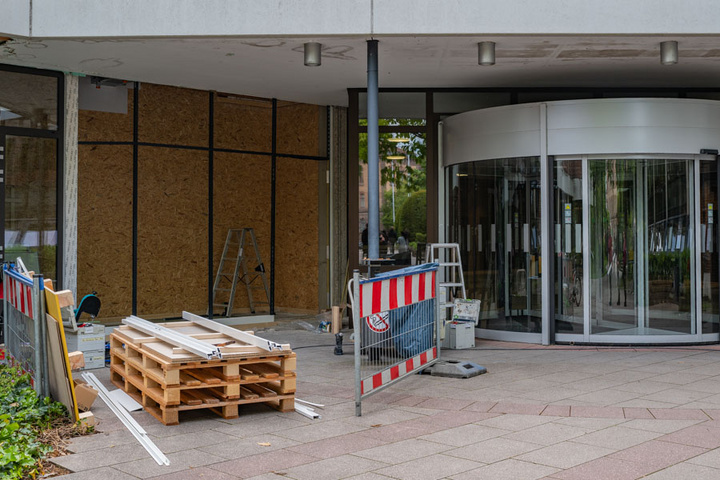 Der Eingang eines Gebäudes zeigt Baustellenmaterialien wie Holzpaletten und Leitern. Ein rotes und weißes Absperrgitter markiert den Bereich, während die Glastüren des Eingangs teilweise geschlossen sind, was auf laufende Renovierungsarbeiten hinweist.