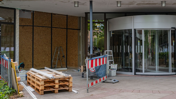 Der Eingang eines Gebäudes zeigt Baustellenmaterialien wie Holzpaletten und Leitern. Ein rotes und weißes Absperrgitter markiert den Bereich, während die Glastüren des Eingangs teilweise geschlossen sind, was auf laufende Renovierungsarbeiten hinweist.
