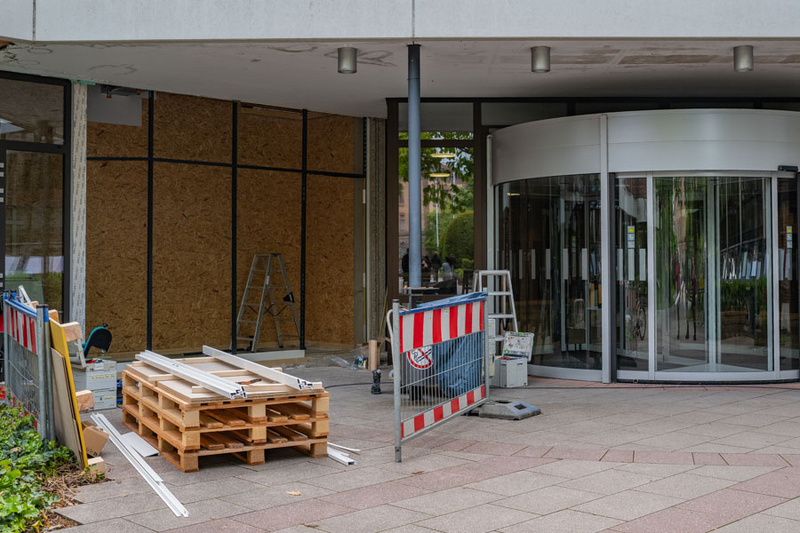 Der Eingang eines Gebäudes zeigt Baustellenmaterialien wie Holzpaletten und Leitern. Ein rotes und weißes Absperrgitter markiert den Bereich, während die Glastüren des Eingangs teilweise geschlossen sind, was auf laufende Renovierungsarbeiten hinweist.