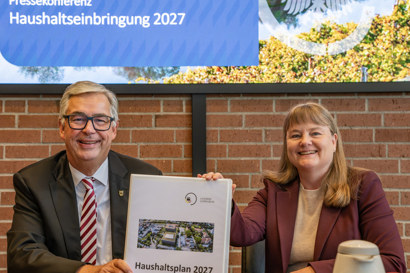 Zwei Personen sitzen an einem Tisch und präsentieren ein Dokument mit dem Titel "Haushaltsplan 2027". Im Hintergrund ist ein Banner mit der Aufschrift "Pressekonferenz Haushalts einbringung 2027" zu sehen. Beide Personen lächeln und sind formell gekleidet. (Text KI generiert)