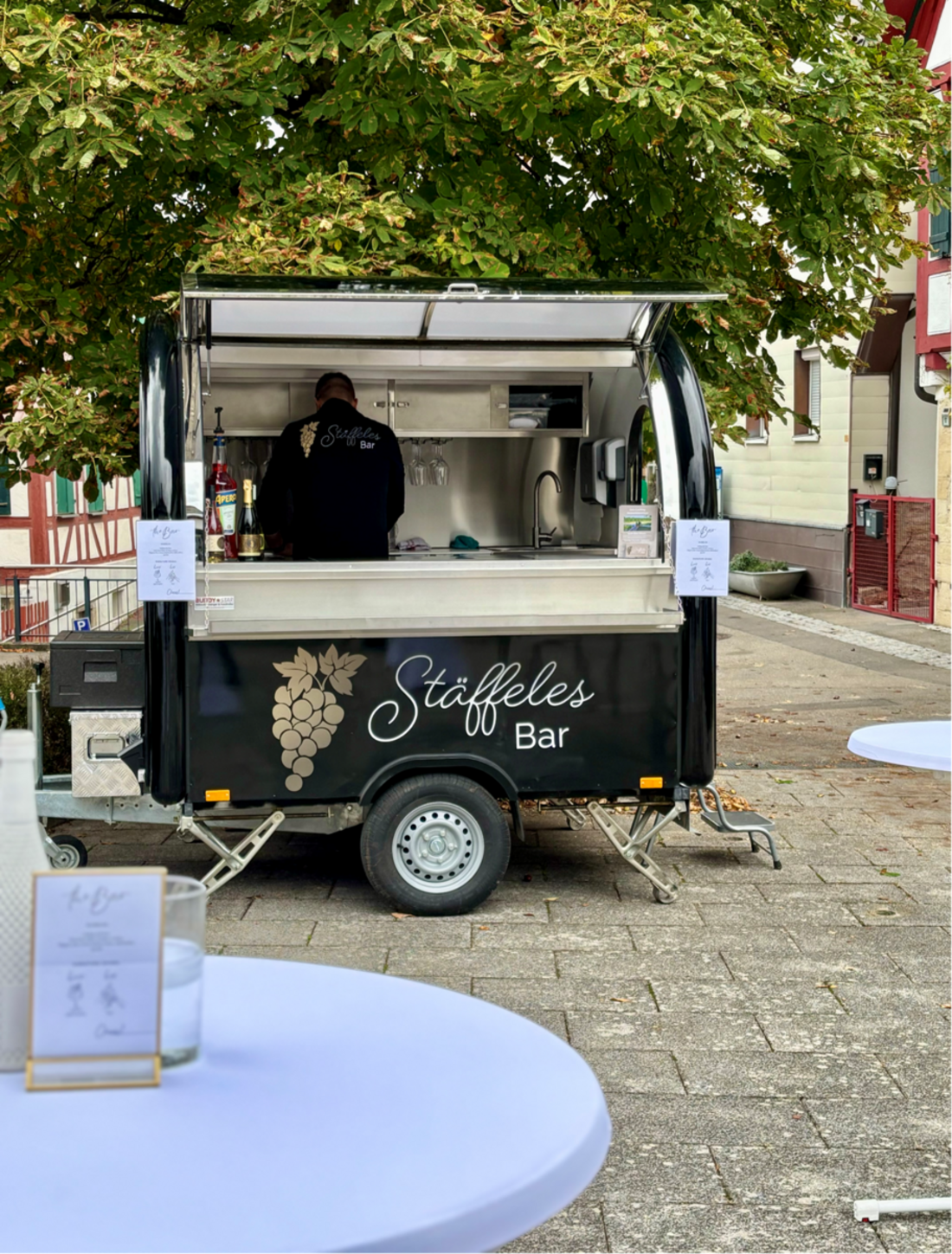 Ein Verkaufswagen mit der Aufschrift „Stäffeles Bar“ steht in einer Fußgängerzone. Ein Mitarbeiter in schwarzem Outfit ist im Inneren des Wagens beschäftigt. Um den Wagen herum befinden sich runde Tische mit blauer Tischdecke.