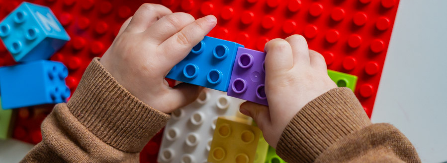 Header Bild: Ein Kind baut mit bunten LEGO-Steinen auf einer roten Basisplatte. Die Hände des Kindes halten verschiedene Steine, während es kreativ an einer Struktur arbeitet. Einige Steine sind in Blau, Lila, Grün und Weiß.