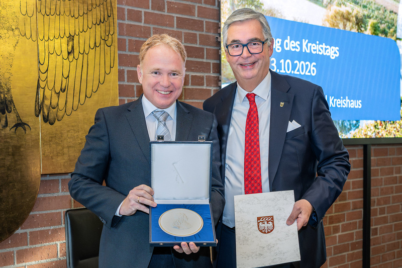 Zwei Männer stehen in einem Raum, lächeln und halten eine Medaille sowie ein Dokument. Der Mann links trägt ein hellblaues Hemd und einen dunklen Anzug, der rechts trägt einen schwarzen Anzug mit einer roten Krawatte. Im Hintergrund ist eine Wand mit einer goldenen Adlerdarstellung zu sehen. (Text KI generiert)