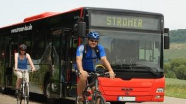 Un autobus rosso con la scritta "STROMER" è fermo su una strada. Due ciclisti, uno dei quali indossa un caschetto blu, stanno passando accanto al bus, mentre un'altra persona si avvicina in bicicletta. Sullo sfondo si vedono colline verdi.