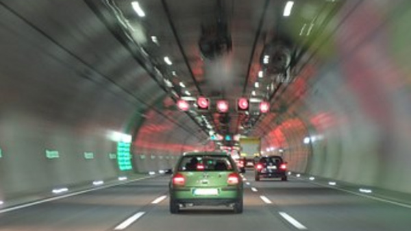 A view inside a tunnel with two vehicles approaching brightly lit traffic signal lights. The lights ahead are red, indicating a stop. The tunnel walls are illuminated with green signs and soft, reflective lighting, creating a sense of motion and depth.