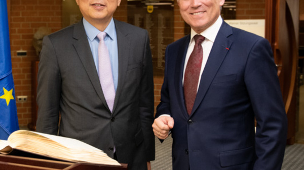 Two men stand smiling beside a large open book on a table. They are flanked by flags: one representing Germany, one for China, and another for the European Union. The setting appears formal, suggesting a diplomatic or official engagement.