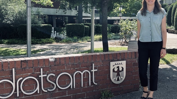 Eine Person steht neben dem Schild des Landratsamts. Das Schild zeigt den Schriftzug "landratsamt" und ein Wappen. Im Hintergrund sind Bäume und ein Gebäude sichtbar. Es ist ein sonniger Tag.