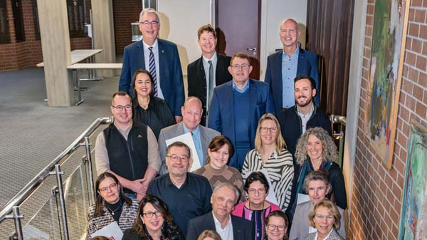 Eine Gruppe von circa 40 Personen steht in einem Treppenhaus und hält Urkunden in der Hand. Sie wirken stolz und lächeln. Im Hintergrund sind kunstvolle Wandbilder und eine moderne Architektur sichtbar.