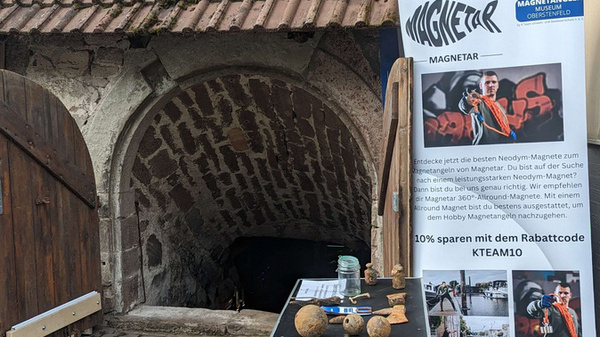 Entrée de cave en maçonnerie avec une porte en bois, devant laquelle sont accrochées des bannières publicitaires concernant le musée Magnetangler d'Oberstenfeld.