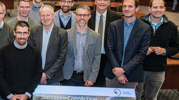 Eine Gruppe von Menschen steht in einem großen Raum. Sie sind in einem semi-formellen Umfang gekleidet und lächeln in die Kamera. Im Vordergrund sind zwei große Banner mit dem Titel „Connecting Connections“ und dem Datum 22.-24.11.2023 in Ludwigsburg zu sehen.