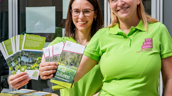 Zwei Frauen stehen an einem Informationstresen und halten Flyer in den Händen. Beide tragen grüne T-Shirts und lächeln. Die Flyer thematisieren das Radfahren und die Erkundung der Natur. Im Hintergrund sind unscharfe informative Elemente zu sehen.