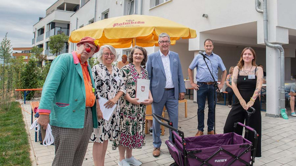 Eine Gruppe von sechs Personen steht im Freien vor einem Gebäude. Eine Person in einem bunten Anzug ist links, während die anderen in formeller Kleidung einen Preis oder eine Auszeichnung halten. Im Hintergrund sieht man eine gelbe Markise.