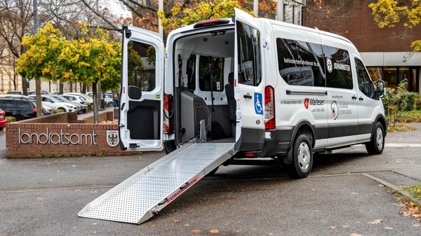 Ein barrierefreier Transporter steht vor einem Landratsamt. Die Tür ist geöffnet, und eine Rampe führt vom Fahrzeug zu dem Boden, um den Zugang für Personen mit Mobilitätseinschränkungen zu erleichtern. Der Transporter ist mit einem Logo versehen.
