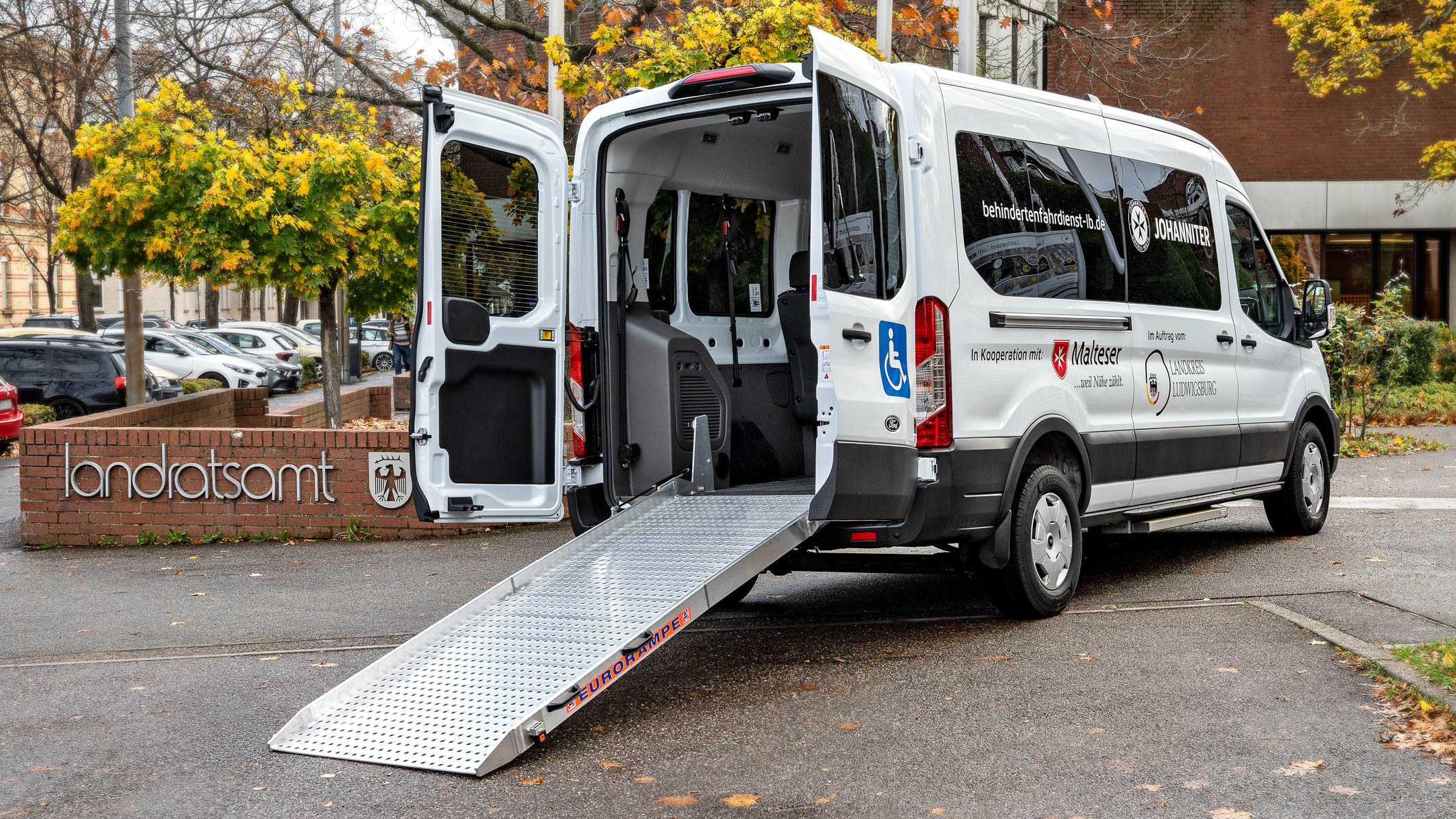 Ein barrierefreier Transporter steht vor einem Landratsamt. Die Tür ist geöffnet, und eine Rampe führt vom Fahrzeug zu dem Boden, um den Zugang für Personen mit Mobilitätseinschränkungen zu erleichtern. Der Transporter ist mit einem Logo versehen.