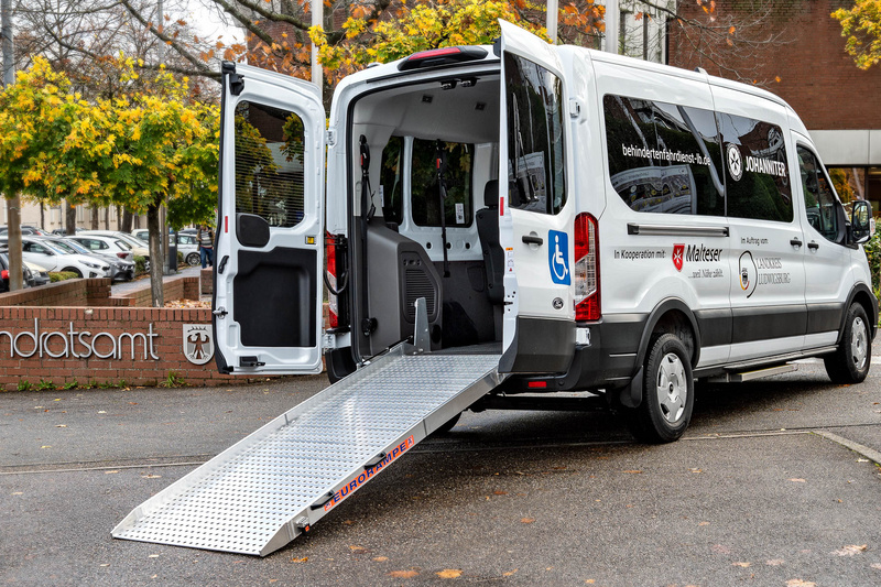Ein barrierefreier Transporter steht vor einem Landratsamt. Die Tür ist geöffnet, und eine Rampe führt vom Fahrzeug zu dem Boden, um den Zugang für Personen mit Mobilitätseinschränkungen zu erleichtern. Der Transporter ist mit einem Logo versehen.