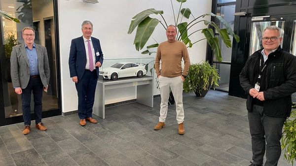 Vier Männer stehen in einem modernen Foyer. Im Hintergrund befindet sich eine große Pflanze und ein Tisch mit einem Modell eines Autos. Die Männer lächeln und tragen formelle Kleidung, während sie miteinander interagieren und den Raum präsentieren. (Text KI generiert)
