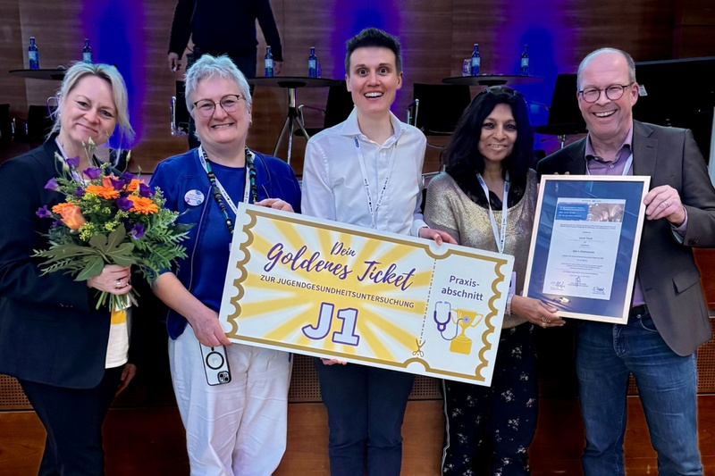Fünf Personen stehen auf einer Bühne, lächeln und halten ein großes Schild mit der Aufschrift „Dein goldenes Ticket“ und dem Zusatz „Preis ausgezeichnet“. Eine Person trägt einen Blumenstrauß. Im Hintergrund sind Stühle und Tische zu sehen.