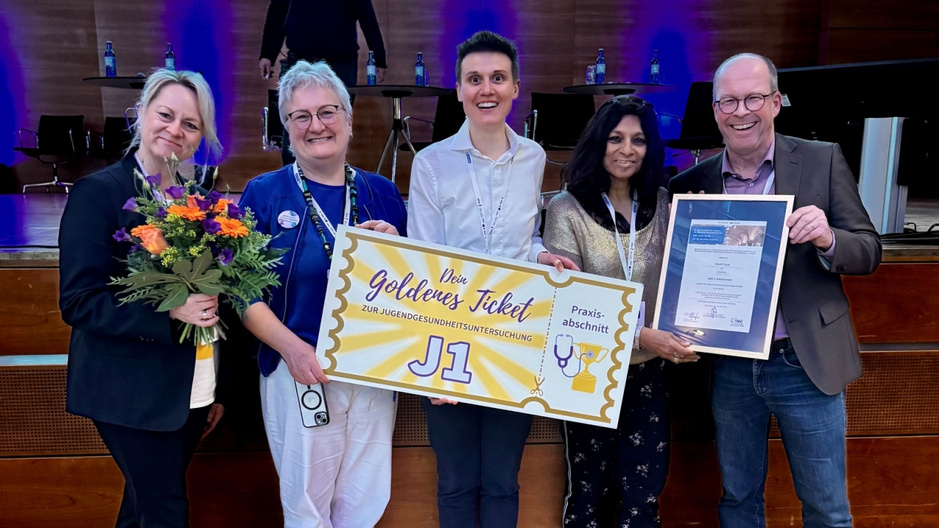 Fünf Personen stehen auf einer Bühne, lächeln und halten ein großes Schild mit der Aufschrift „Dein goldenes Ticket“ und dem Zusatz „Preis ausgezeichnet“. Eine Person trägt einen Blumenstrauß. Im Hintergrund sind Stühle und Tische zu sehen.