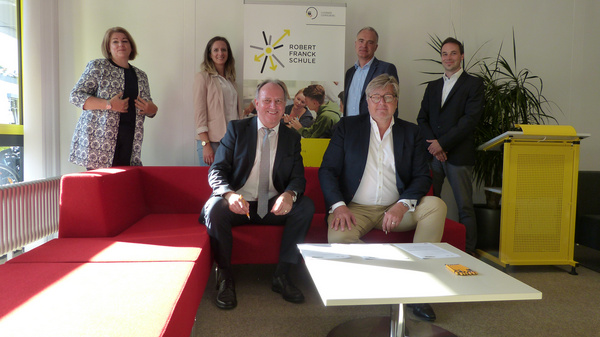 Un groupe de six personnes se tient dans un espace intérieur lumineux, avec une banquette rouge et une table basse au centre. Un panneau avec le logo de la Robert Franck Schule est visible en arrière-plan. Les individus, habillés de manière professionnelle, semblent discuter ou poser pour la photo.