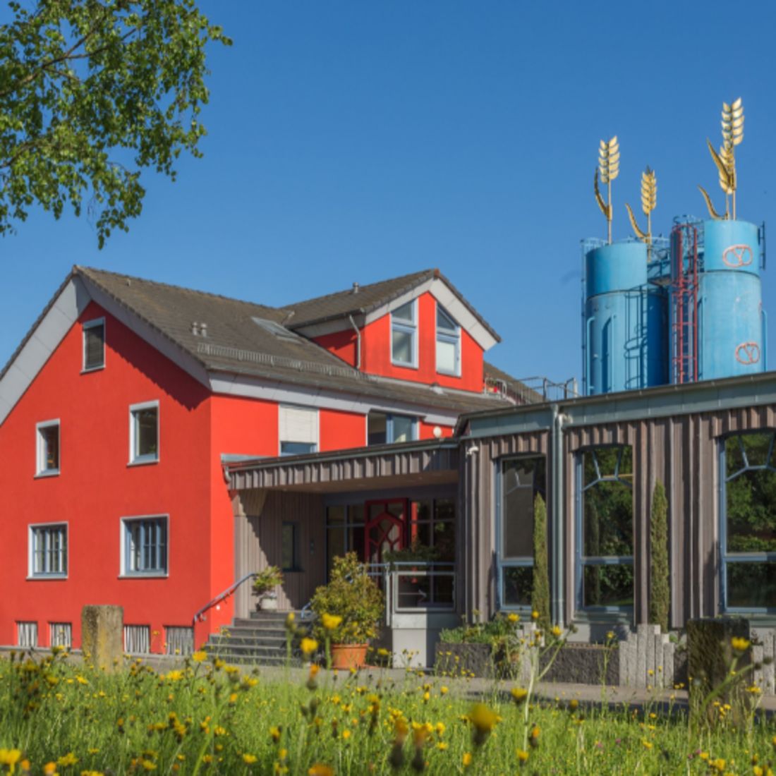 Das Bild zeigt ein rotes, mehrstöckiges Haus mit Spitzdach. Rechts ist ein einstöckiger, brauner Anbau mit Flachdach, auf dem zwei blaue Silos sind. Die Silos sind mit Brezeln bemalt und goldenen Skulpturen in der Form von Getreideähren gekrönt.