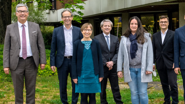 Eine Gruppe von acht Personen steht draußen in einer Gartenlandschaft. Sie sind in formeller Kleidung gekleidet und lächeln in die Kamera. Die Szene vermittelt ein Gefühl von Zusammengehörigkeit und Professionalität.