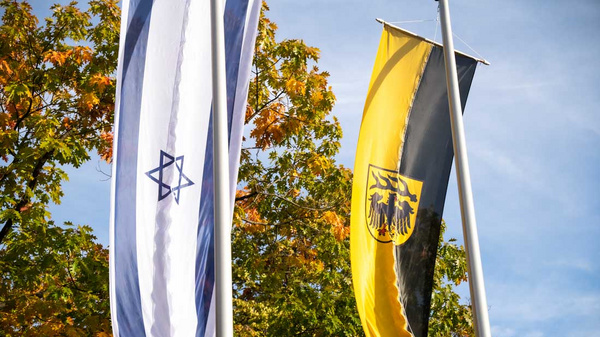 Zwei Fahnen wehen im Wind: eine mit dem Davidstern und eine in Schwarz-Gelb, die das Wappen zeigt. Im Hintergrund sind bunte Herbstblätter zu sehen.