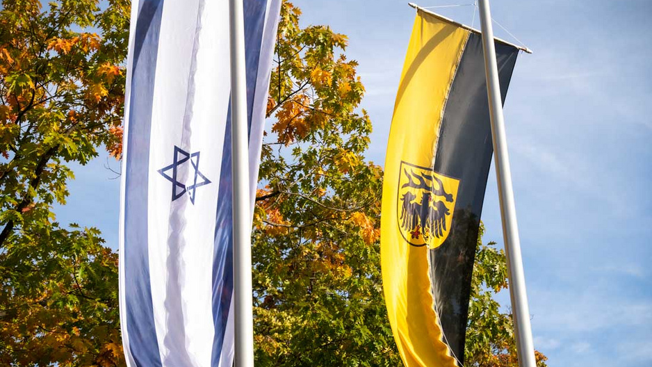 Zwei Fahnen wehen im Wind: eine mit dem Davidstern und eine in Schwarz-Gelb, die das Wappen zeigt. Im Hintergrund sind bunte Herbstblätter zu sehen.