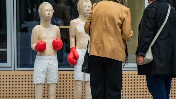 Zwei Skulpturen von Boxern stehen in einem Ausstellungsbereich. Beide Figuren tragen rote Boxhandschuhe und sind in weißen Shorts gekleidet. Zwei Frauen, eine mit langen grauen Haaren und eine mit kurzen braunen Haaren, betrachten die Skulpturen interessiert.