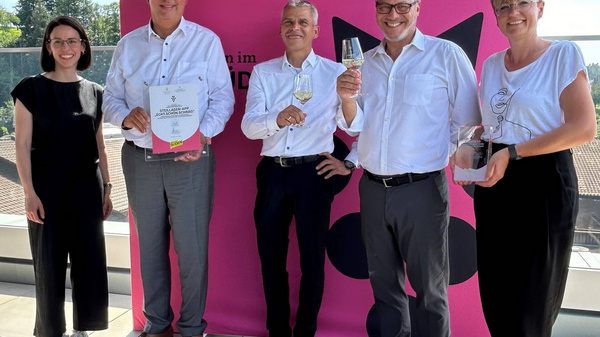Cinq personnes se tiennent debout sur une terrasse, souriant. Elles portent des vêtements professionnels. Deux d'entre elles tiennent des trophées, tandis qu'une autre montre un certificat. En arrière-plan, un paysage verdoyant est visible sous un ciel ensoleillé. Un fond rose avec le texte "Wir sind Wein" est présent.