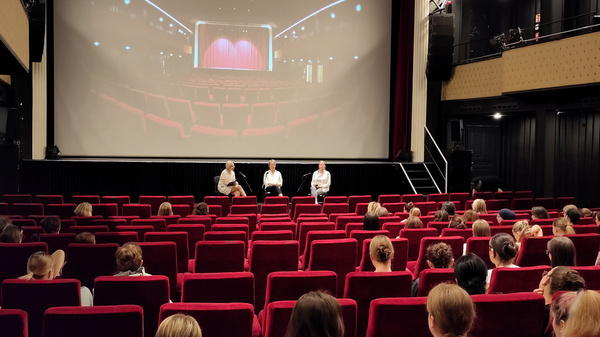 Un auditorium di un cinema con posti a sedere rossi. Sul palco, tre persone parlano di fronte a un grande schermo. In sala, un pubblico composto principalmente da giovani ascolta attentamente. L'atmosfera è di attenzione e partecipazione.