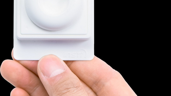 Une main tient un objet blanc de forme carrée avec un bouton circulaire en relief au centre. L'arrière-plan est noir, mettant en valeur l'objet. C'est un appareil électronique, probablement un capteur ou un contrôle, mais aucune inscription n'est visible.