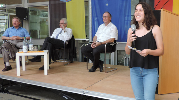 A young woman stands on stage speaking into a microphone, while three seated older men listen. The backdrop features flags of Germany and the European Union, suggesting the event focuses on civic engagement or political discussion. A wooden podium is present on stage.