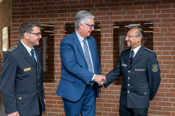 Drei Männer in Uniform stehen in einem Innenraum mit Backsteinwand. Zwei der Männer haben lächelnde Gesichter und schütteln die Hände des dritten Mannes, der im Anzug gekleidet ist. Im Hintergrund sind Wandschilder angebracht.