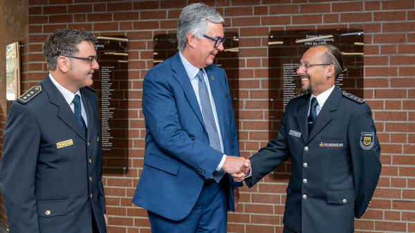 Drei Männer in Uniform stehen in einem Innenraum mit Backsteinwand. Zwei der Männer haben lächelnde Gesichter und schütteln die Hände des dritten Mannes, der im Anzug gekleidet ist. Im Hintergrund sind Wandschilder angebracht.