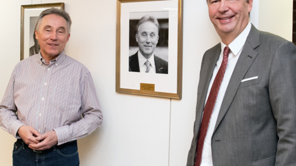 Two men stand next to a gallery wall filled with framed portraits. One man, wearing a striped shirt, smiles while the other, dressed in a suit, stands beside him. The setting appears to be a modern indoor space with seating.