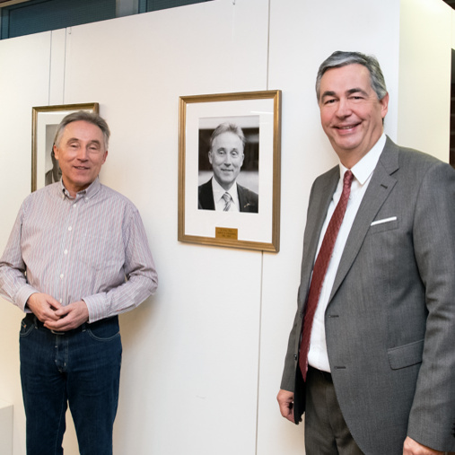 Zwei Männer stehen entspannt vor einer Wand mit gerahmten Porträtfotos. Die Männer lächeln und scheinen sich zu unterhalten. Im Hintergrund sind mehrere Portraits in Rahmen zu sehen, die auf eine Ausstellung hinweisen.