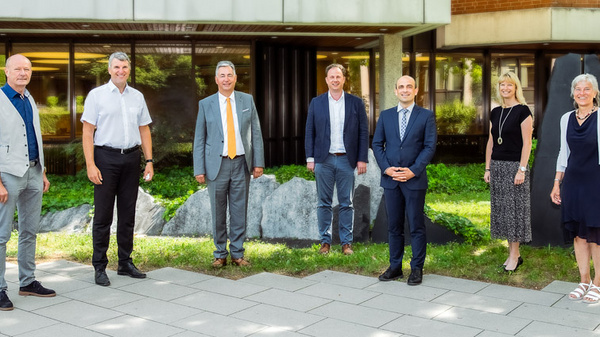 A group of seven professionals stands together outdoors near a landscaped area. They are dressed in business attire and appear to be engaged in a collaborative or formal setting. The background features greenery and architectural elements.