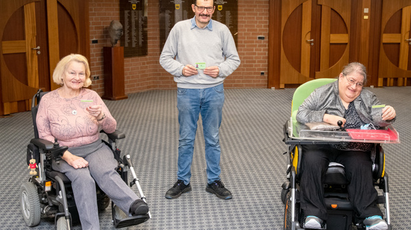 A man stands between two women in wheelchairs, all engaging in conversation. The woman on the left has blonde hair and smiles while holding a cup. The woman on the right, with short hair, is also smiling and has a tray in front of her. The setting is indoors with wooden accents.