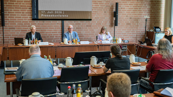 Eine Pressekonferenz am 17. Juli 2026 wird abgehalten. An einem Tisch sitzen drei Personen, ein Mann und eine Frau in professioneller Kleidung. Im Publikum sind mehrere Personen sichtbar, die auf die Redner blicken. Auf dem Tisch stehen Getränke und Snacks.