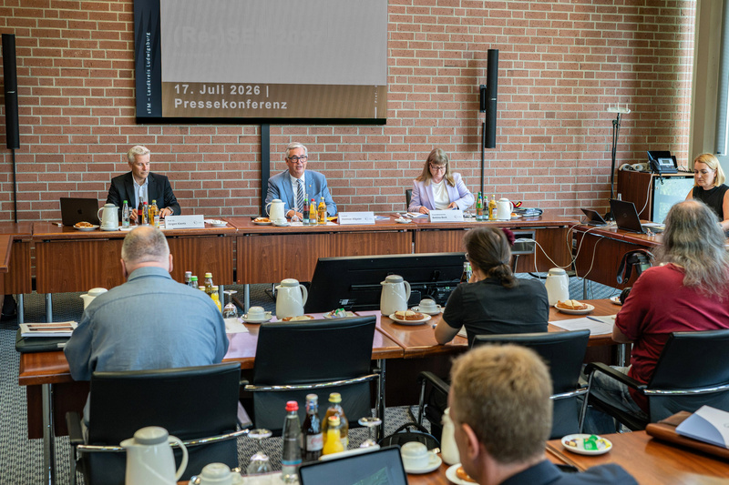 Eine Pressekonferenz am 17. Juli 2026 wird abgehalten. An einem Tisch sitzen drei Personen, ein Mann und eine Frau in professioneller Kleidung. Im Publikum sind mehrere Personen sichtbar, die auf die Redner blicken. Auf dem Tisch stehen Getränke und Snacks.