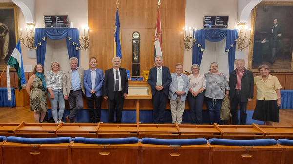 Eine Gruppe von zwölf Personen steht in einem Saal mit Holzmöbeln und Flaggen. Im Hintergrund sind große Gemälde an den Wänden sichtbar. Die Personen lächeln und posieren zusammen, während sie sich für ein Foto aufstellen.
