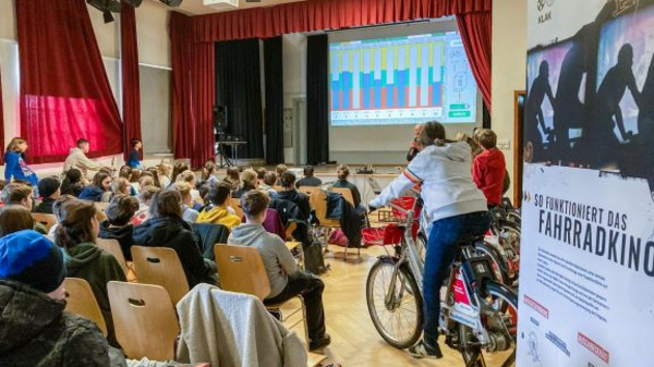 Eine Schulveranstaltung zeigt ein Publikum von Schülern, das einem Vortrag folgt. Auf der Bühne wird eine Präsentation mit bunten Diagrammen angezeigt. Links steht ein Plakat mit Informationen zum Thema Fahrradkino, während eine Person auf einem Fahrrad sitzt und an der Veranstaltung teilnimmt.